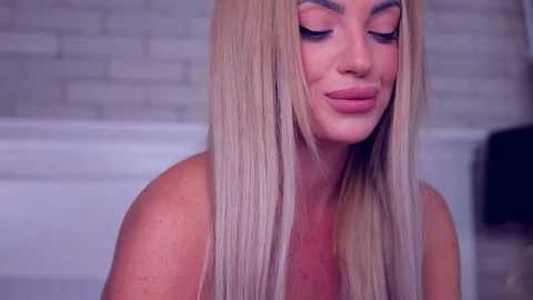 Media: A video of a topless blonde woman with long, straight hair, wearing subtle makeup and a closed-mouth smile. She stands in front of a white tiled wall, with a blurred black object in the background.