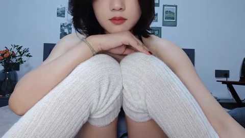 Media: Video of an Asian woman with fair skin, wearing white knee-high socks and red lipstick, sitting cross-legged on a bed with a floral arrangement in the background.