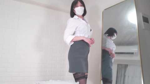 Media: Video of an Asian woman with a white mask, wearing a white blouse, grey pencil skirt, and black lace stockings, standing in front of a mirror in a white brick-walled room.