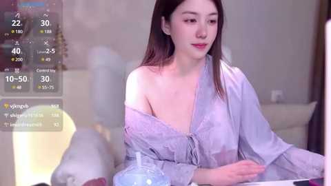 Media: Video of a young Asian woman with straight, dark hair, wearing a light grey off-shoulder blouse, sitting at a table in a softly lit room. Background includes a digital weather display with temperatures and icons.