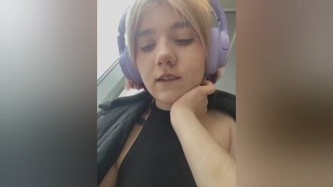 Media: A close-up video of a young woman with light skin, blonde hair, and large purple headphones, resting her chin on her hand. She wears a black top, and the background is blurred.