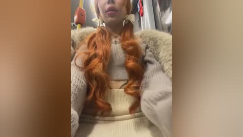 Media: Video of a woman with long, wavy, red hair, wearing a cream-colored, fur-trimmed sweater, seated indoors, with a blurred background featuring a hanging red lantern and white clothing.