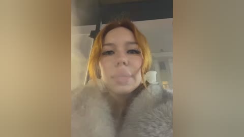 Media: A video of a young woman with fair skin and orange hair, wearing a fluffy, light-colored coat, blowing a bubble, taken indoors with blurred background.