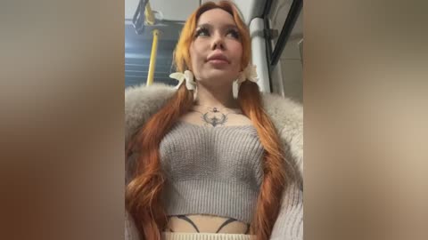 Media: A video of a young woman with long, wavy red hair, wearing a grey ribbed crop top, white fur coat, and large white flower earrings. She stands in a subway car with yellow poles in the background.