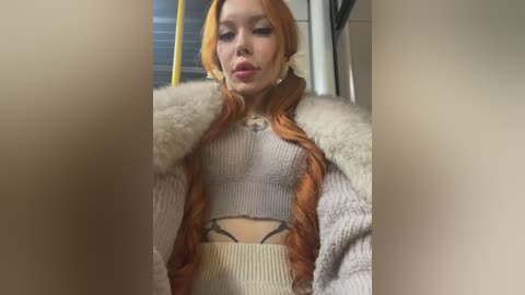 Media: Video of a young woman with long, vibrant orange hair in braids, wearing a cream knitted sweater, beige skirt, and white fur coat, standing indoors near a door.