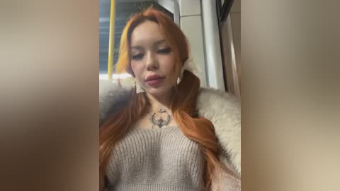 Media: Video of a young woman with fair skin, long red hair, wearing a beige knit sweater, sitting indoors. She has a small tattoo on her chest and is looking down.