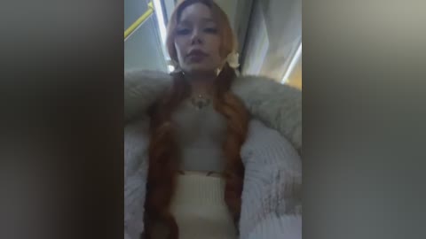 Media: Video of a young woman with light skin, long blonde hair in twin braids, wearing a beige sweater and a white fur coat, seated in a dimly lit room with a yellow wall and a white blanket.