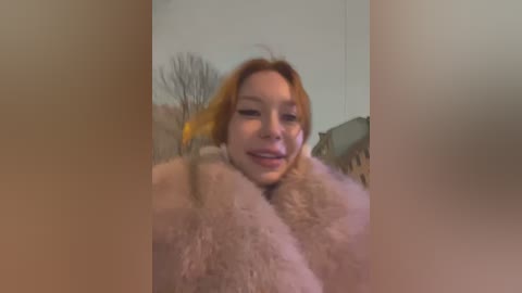 Media: A blurry, grainy video of a smiling woman with fair skin and shoulder-length blonde hair, wrapped in a fluffy, beige blanket, standing indoors with a bare tree visible outside.