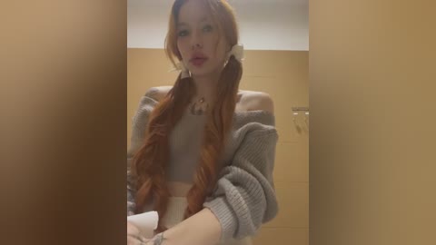 Media: Video of a young woman with long, wavy red hair in pigtails, wearing a gray off-shoulder sweater and white bralette, standing in a beige-tiled bathroom.