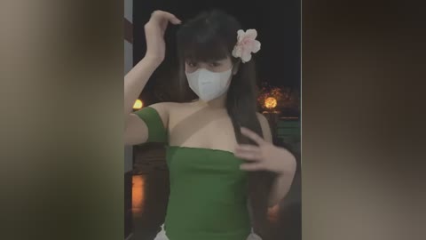 Media: A video of an East Asian woman with long black hair and a pink flower in her hair, wearing a green off-shoulder top and white face mask, standing in a dimly lit room with candles and a fireplace.