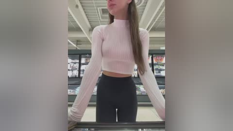 Media: Video of a slender, light-skinned woman with long brown hair wearing a white ribbed crop top and high-waisted black leggings. She stands in a modern, brightly lit supermarket aisle with shelves filled with products in the background.