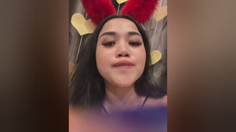 Media: Video of a young woman with long black hair, wearing red and yellow heart-shaped bunny ears, lying on a bed with a gray blanket. She has light skin, a slight smile, and is topless.