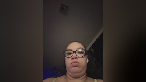 Video of a woman with a large head, fair skin, and glasses, sitting indoors with a dimly lit background and a small vent on the ceiling.