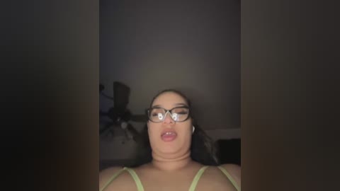 Video of a light-skinned woman with dark hair, wearing glasses, a green bra, and an open-mouthed expression. The background is dimly lit, featuring a fan and a chair.