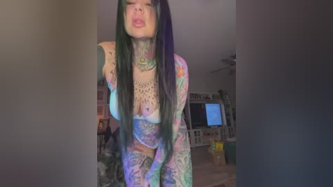 Media: Video of a tattooed woman with long dark hair, wearing a colorful, see-through bodysuit, bending forward in a living room with a TV, bookshelf, and dim lighting.