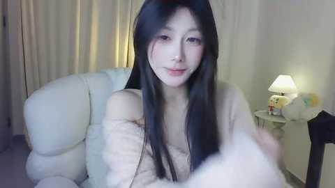 Media: A video of an East Asian woman with long black hair, fair skin, and a soft smile, wearing a beige off-shoulder sweater, sitting on a white couch in a softly lit room.