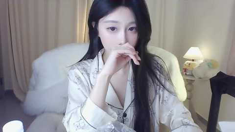 Media: Video of an Asian woman with long black hair, wearing a white, patterned robe, seated in a dimly lit bedroom with a lamp, plush toys, and beige curtains.