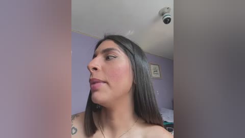 Media: Video of a Latina woman with straight dark hair, wearing minimal makeup, and a silver necklace. She has a tattoo on her left shoulder. The background shows a purple wall with a ceiling-mounted camera and a framed picture.