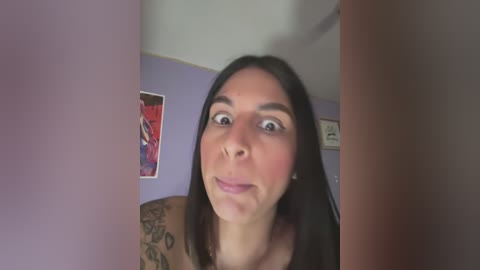 Media: A video of a Latina woman with long black hair and a tattoo sleeve, wearing a black top, looking surprised. The background features a purple wall with framed artwork.