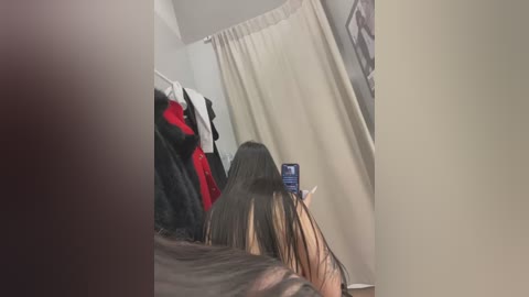 Media: Video of a woman with long, straight brown hair, lying on a bed, taking a selfie in a dimly lit room with beige curtains and clothes hanging on a rack.