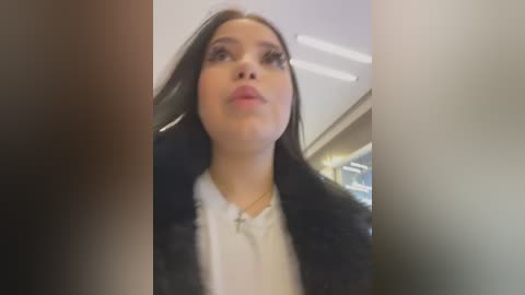 Media: Video of a young woman with long dark hair, wearing a black fur coat over a white shirt, looking upward with a neutral expression, blurred background.