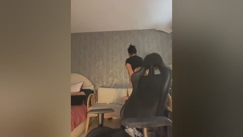 Media: A video captures a young Asian woman with a dark bun, wearing a black top, sitting on a black gaming chair, facing a beige couch with a red blanket and a white pillow in a dimly lit room with a textured gray wall.