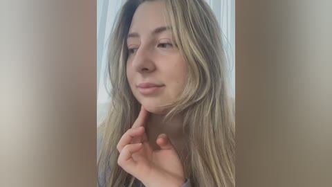 Media: Video of a young woman with light skin, long blonde hair, and a contemplative expression, touching her chin. She wears a gray shirt, and the background is blurred, focusing on her face.