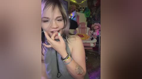 Media: A video of a young woman with light skin, short blonde hair, and a sleeve tattoo, wearing a grey tank top, dancing and playing with a cigarette in a dimly lit, colorful room with gaming chairs and a green plush toy.