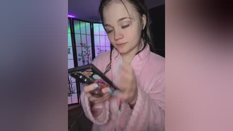 Media: Video of a young woman with wet hair, wearing a pink robe, engrossed in a smartphone, indoors with dim lighting and a decorative screen in the background.