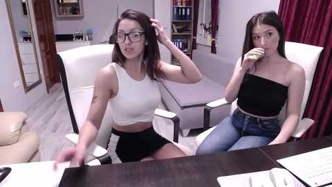 Media: Video of two women in a modern office. One with glasses, white crop top, black shorts, seated at desk. Other, brunette, in black strapless top, jeans, eating a sandwich, seated in white chair.