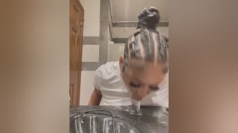 Media: Video of a young woman with dark braided hair, wearing a white shirt, drinking from a sink in a kitchen with beige walls and wooden door.