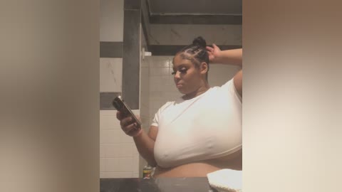 Media: A video of a pregnant Black woman with dark skin, large breasts, and braided hair, wearing a white crop top and gray pants, taking a selfie in a bathroom with tiled walls and a visible ceiling beam.