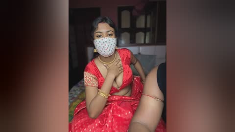 Media: Video of a South Asian woman in a red saree, wearing a face mask, sitting on a bed. The background shows a dimly lit room with shelves and a red wall.