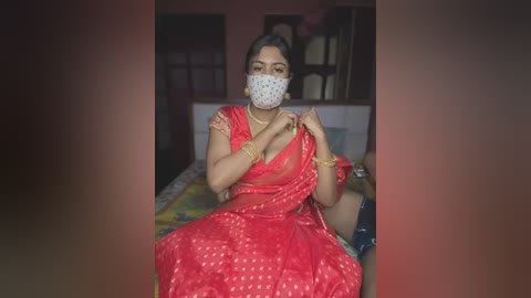 Media: Video of a South Asian woman in a red saree, adjusting her mask, seated on a bed in a dimly lit room with a patterned bedspread.