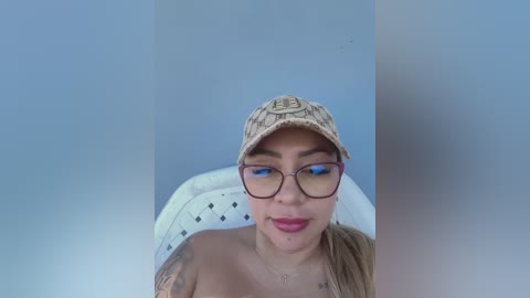 Media: Video of a young Latina woman with light brown skin, wearing glasses, a beige baseball cap, and a white top, sitting in a white chair with diamond-patterned fabric.