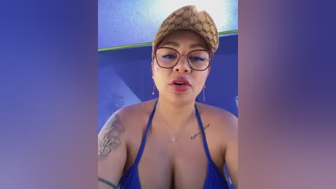 Media: Video of a young, light-skinned woman with medium-length hair and glasses, wearing a blue bikini and a straw hat, with tattoos on her arms, in a blue-tiled room.