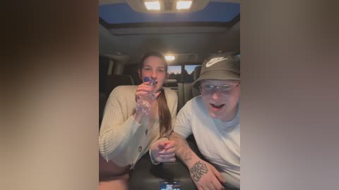 Media: Video of two people, a woman and a man, drinking water in a dimly lit car. Woman in cream cardigan, man in glasses and baseball cap.