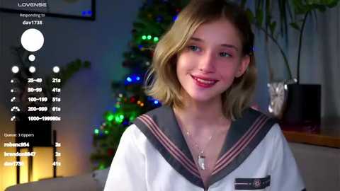 Media: Video of a young woman with shoulder-length blonde hair, wearing a sailor-style school uniform, smiling in a cozy indoor setting with festive decorations and a digital display showing user statistics.