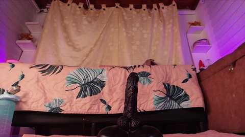 A video of a cozy bedroom with a pink quilt featuring green palm leaves, a black dildo resting on the bed, and a beige curtain with white floral patterns.