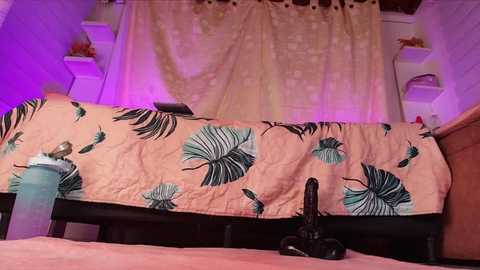 Video of a pink bedspread with green leaf patterns, a black dildo on the bed, and purple lighting in a cozy, dimly-lit bedroom.