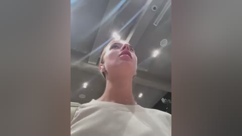 Media: Video of a woman with blonde hair, wearing a white shirt, gazing upward with a neutral expression, captured from a low angle in a modern, dimly lit room with recessed lights and industrial-style ceiling.