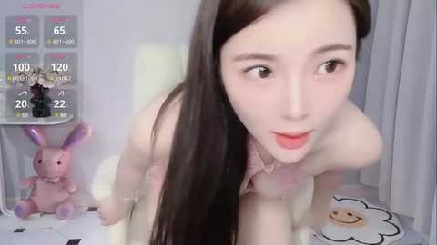 Media: Video of a young Asian woman with long black hair, fair skin, and light pink lipstick, posing in a room with a pink bunny toy, white wall, and a TV displaying live stream stats.