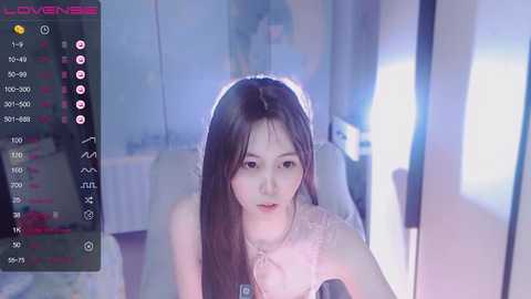 Media: A video of a young East Asian woman with long, straight black hair, wearing a light pink lace top, taken from a webcam, set against a softly lit, dimly-lit room with minimal decor.