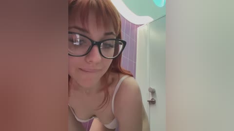 Video of a young woman with light skin, red hair, and black-rimmed glasses, wearing a white bra, smiling slightly. Background shows a bathroom with lavender wallpaper and a white door.