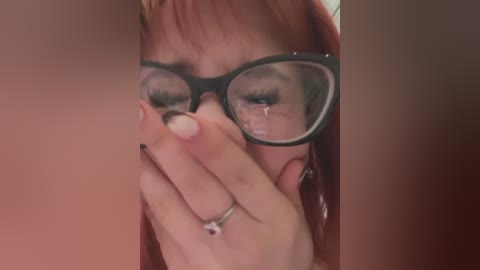 Media: Video of a tearful woman with red hair, wearing black-rimmed glasses and a ring, covering her mouth with her hand.