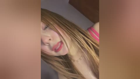 Media: Video of a close-up of a young Caucasian woman with light skin and long blonde hair, wearing a pink top and a simple necklace, taken indoors with soft lighting.