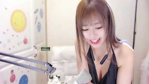 Media: A video of a smiling East Asian woman with straight brown hair, wearing a black halter top, recording herself with a microphone and stand in a brightly lit, decorated room.