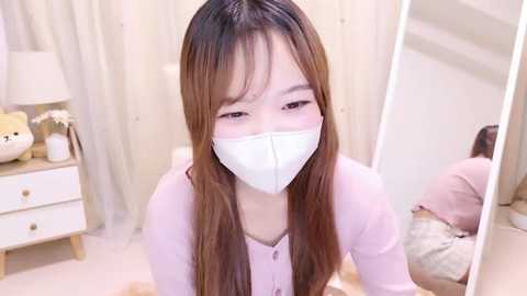 Media: Video of a young Asian woman with long brown hair and fair skin, wearing a pink cardigan and white mask, kneeling in a cozy, minimalist bedroom with cream curtains and a beige bed.