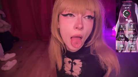 Media: Video of a young woman with pale skin, blonde hair, and dark makeup, sticking out her tongue, wearing a black, ripped top. Background shows a dimly lit room with purple lighting.