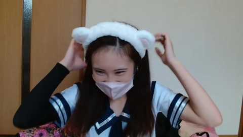 Media: Video of an Asian woman with long brown hair, wearing a white face mask and a black-and-white sailor uniform with a blue tie, playfully adjusting white bear ear headbands.